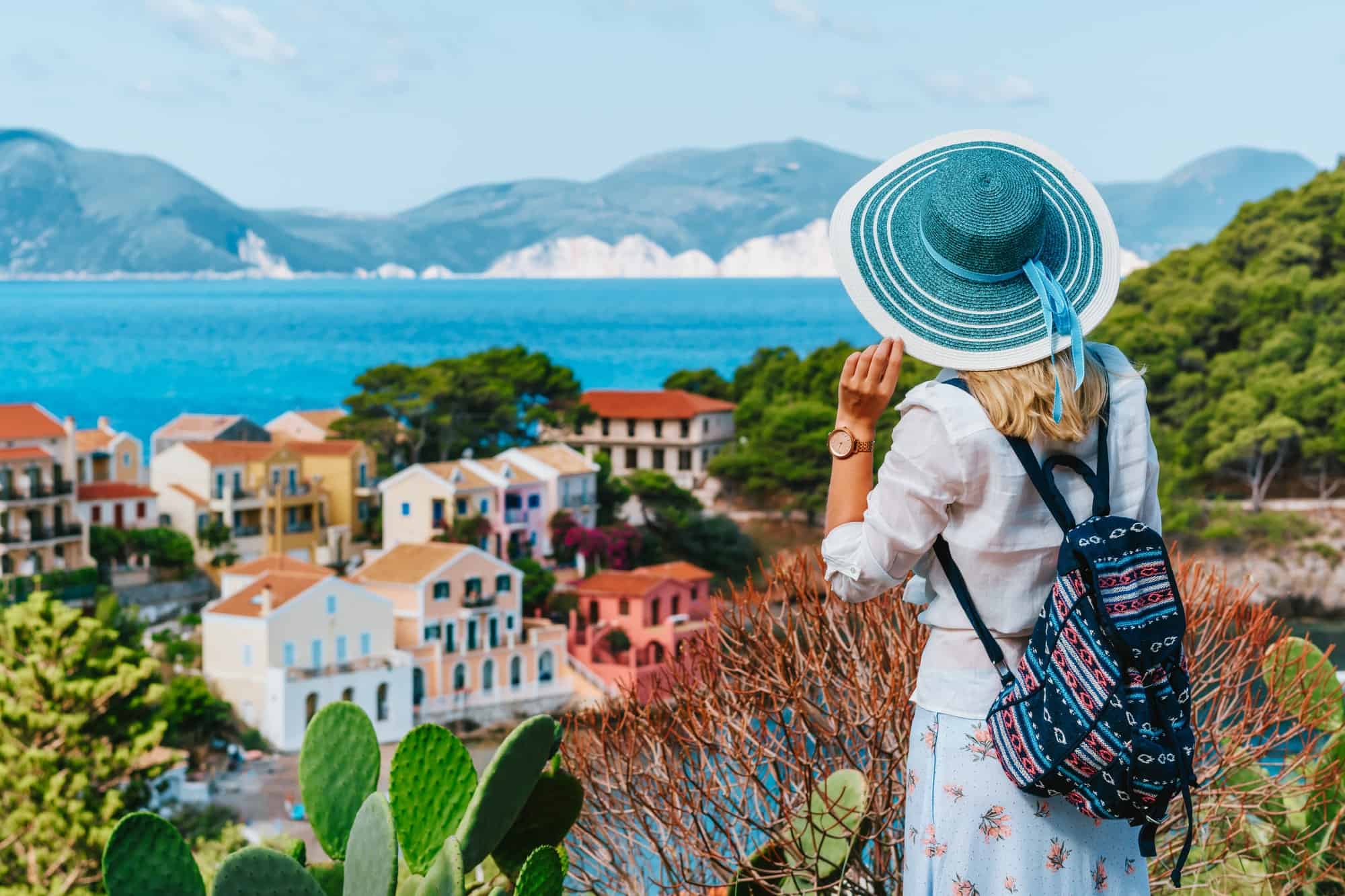 Tourist female with blue sun hat and travel backpack enjoying greek vivid colored Assos town